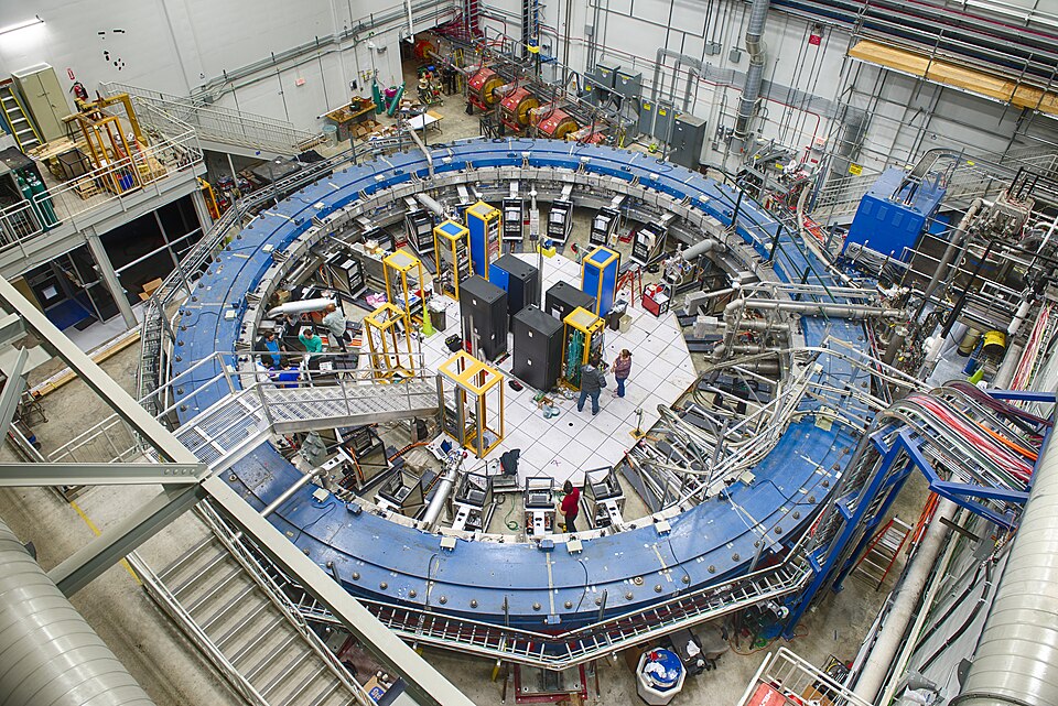 Overhead view of the Muon g-2 storage ring at Fermilab, a large blue circular electromagnet in its detector hall with researchers and equipment inside the ring.
